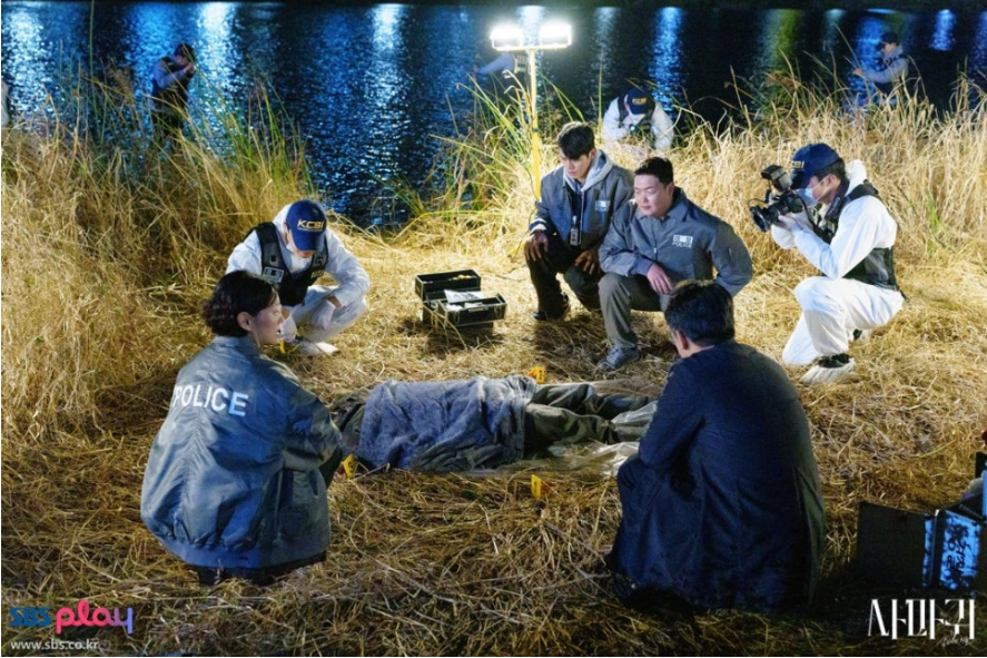 밤 시간, 물가 갈대밭에서 경찰과 국과수 요원들이 변사체를 둘러싸고 현장을 조사하는 모습입니다. 중앙에 시신이 담요로 덮여 있으며, 주변에는 경찰(POLICE)과 과학수사요원(KCSI) 복장의 사람들이 사건의 단서를 찾는 긴박한 분위기를 보여줍니다. 밤 시간, 물가 갈대밭에서 경찰과 국과수 요원들이 변사체를 둘러싸고 현장을 조사하는 모습입니다. 중앙에 시신이 담요로 덮여 있으며, 주변에는 경찰(POLICE)과 과학수사요원(KCSI) 복장의 사람들이 사건의 단서를 찾는 긴박한 분위기를 보여줍니다.