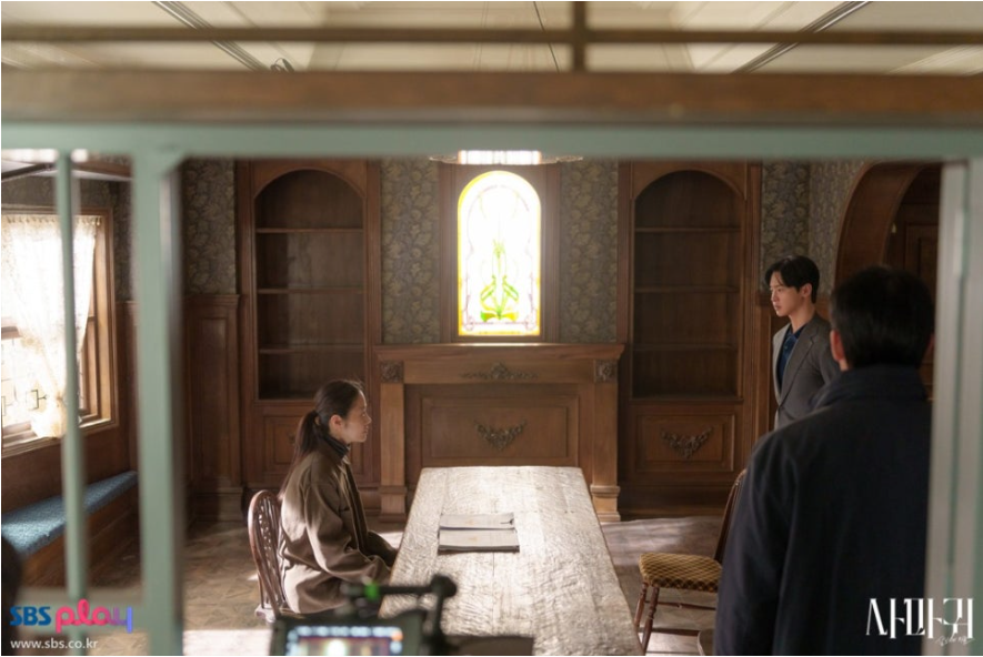 고전적인 분위기의 실내에서 세 인물이 긴 탁자를 사이에 두고 마주하고 있습니다. 고현정으로 보이는 여성 캐릭터는 왼쪽에 앉아 있고, 장동윤으로 보이는 남성 캐릭터는 오른쪽에 서 있으며, 다른 중년 남성이 뒷모습으로 보입니다. 창문의 스테인드글라스가 독특한 분위기를 더합니다. 고전적인 분위기의 실내에서 세 인물이 긴 탁자를 사이에 두고 마주하고 있습니다. 고현정으로 보이는 여성 캐릭터는 왼쪽에 앉아 있고, 장동윤으로 보이는 남성 캐릭터는 오른쪽에 서 있으며, 다른 중년 남성이 뒷모습으로 보입니다. 창문의 스테인드글라스가 독특한 분위기를 더합니다.